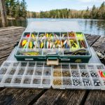A neatly organized fishing tackle box filled with colorful lures, hooks, sinkers, and swivels laid open on a wooden dock